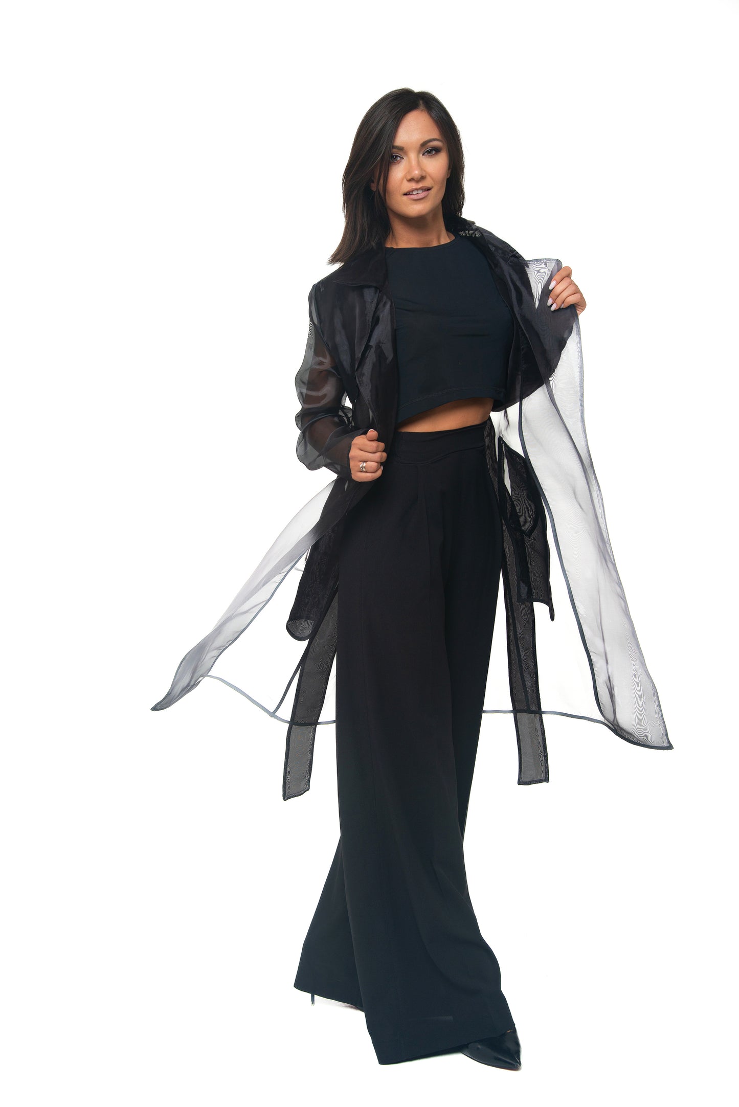 Woman wearing a black outfit with a long, sheer black trench on a white background
